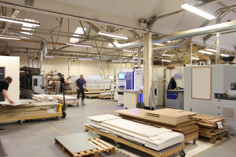 Factory Floor of The Kitchen Depot Glasgow Kitchen Manufacturing Plant. There are kitchen boards, CNC cutting machines and 2 workers moving in the background. The space is busy and a shot of a working factory
